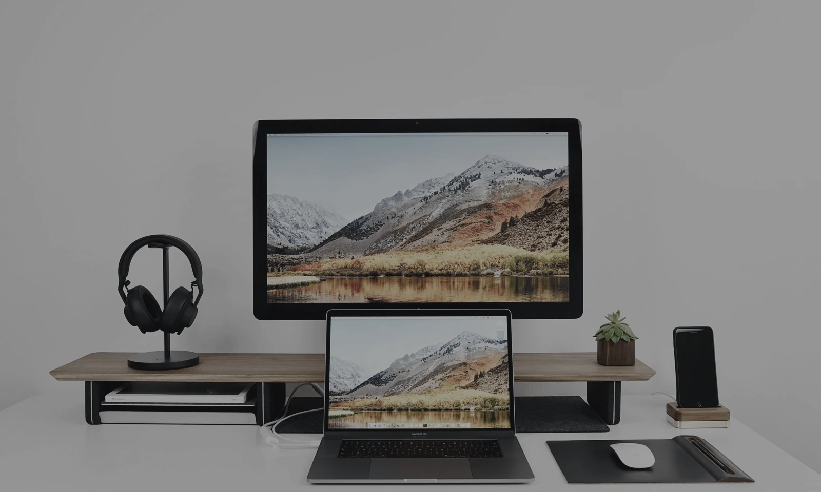 Minimal desk setup with a monitor and laptop showing a mountain landscape plus headphones, phone and mouse